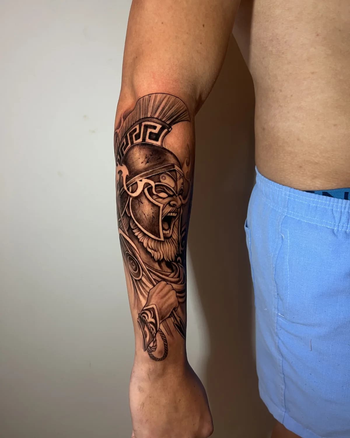 Tatuagem de guerreiro espartano com elmo e grega, preto e cinza no antebraço