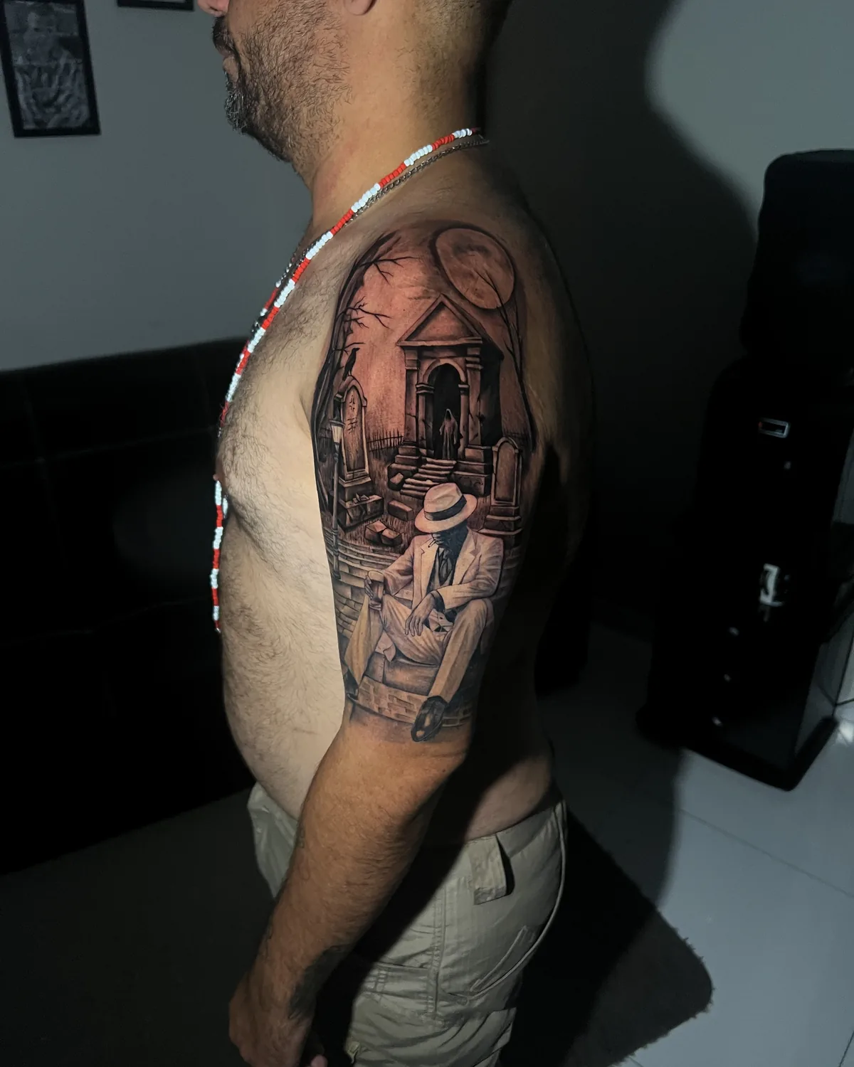 Tatuagem realista de Zé Pilintra sentado, de terno branco e chapéu, com casario e lua ao fundo, no braço